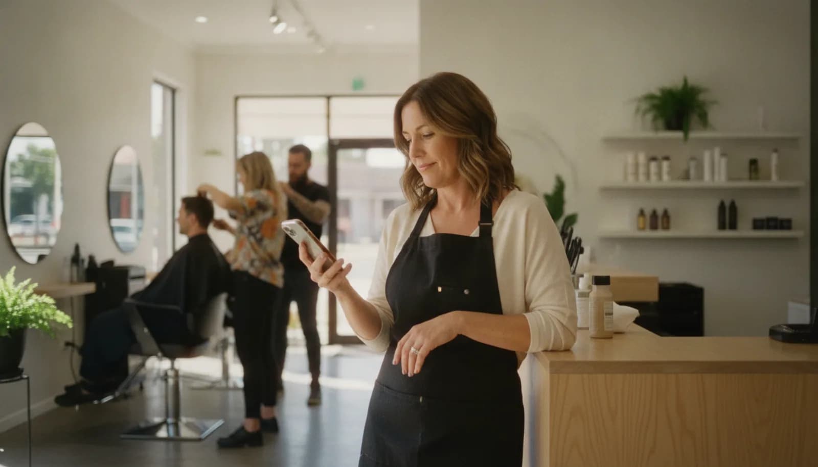 A salon owner checking her phone at the front counter while stylists work behind her